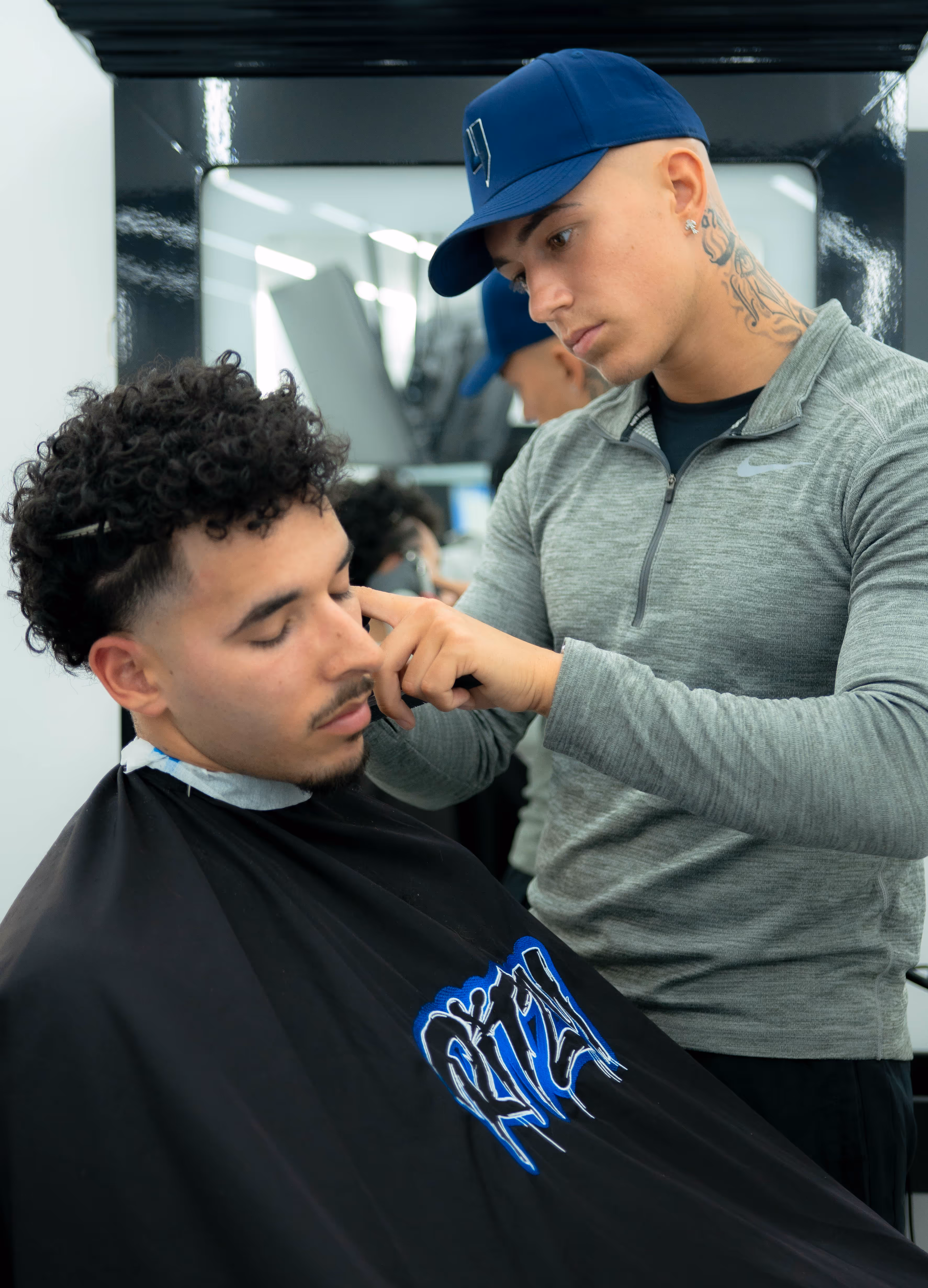 Barber wearing a blue cap and gray Nike top carefully trimming hair on a client's temple in a modern barbershop.
