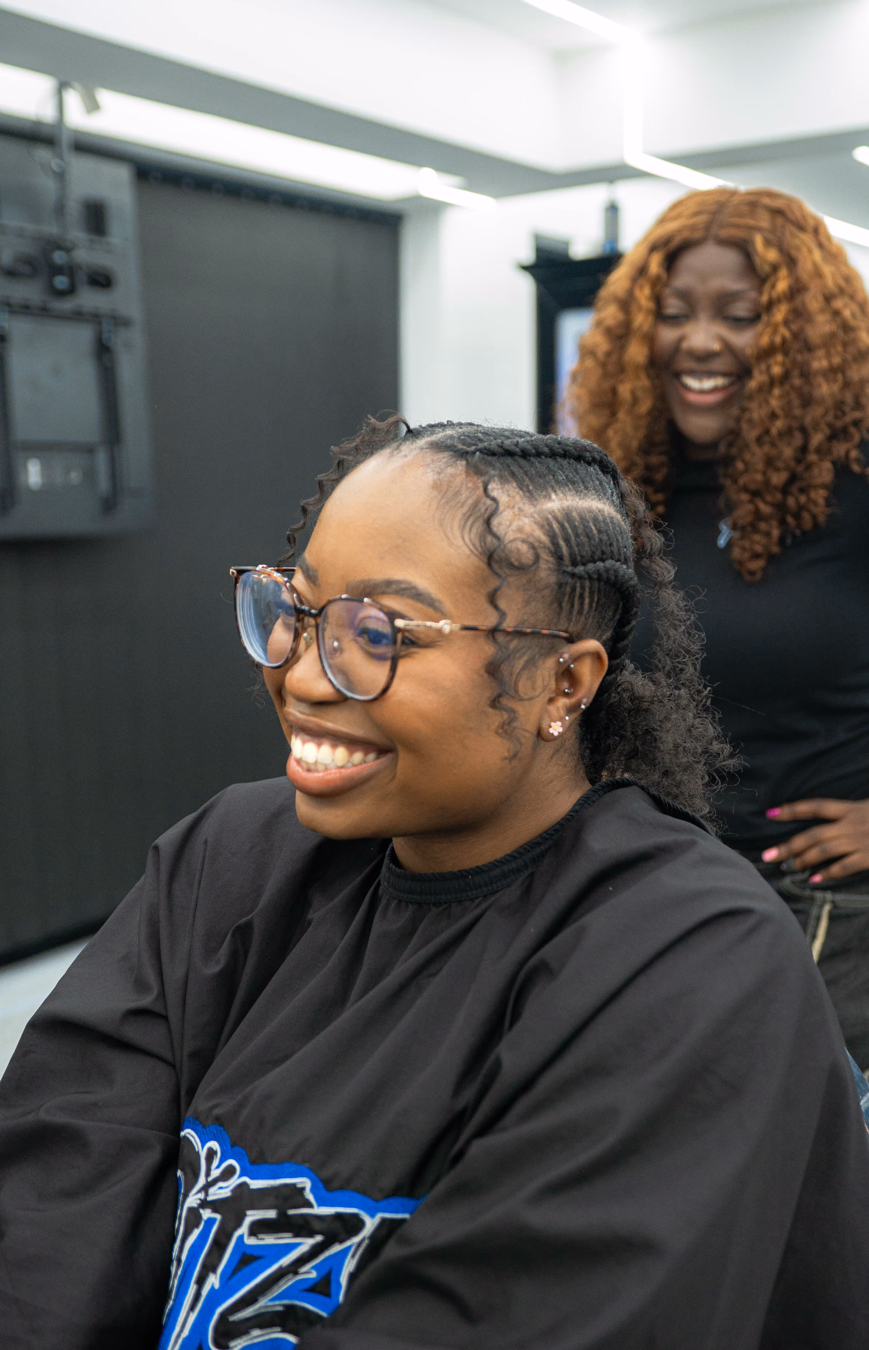 Smiling woman with cornrows and glasses sitting in a salon chair with another woman smiling in the background.