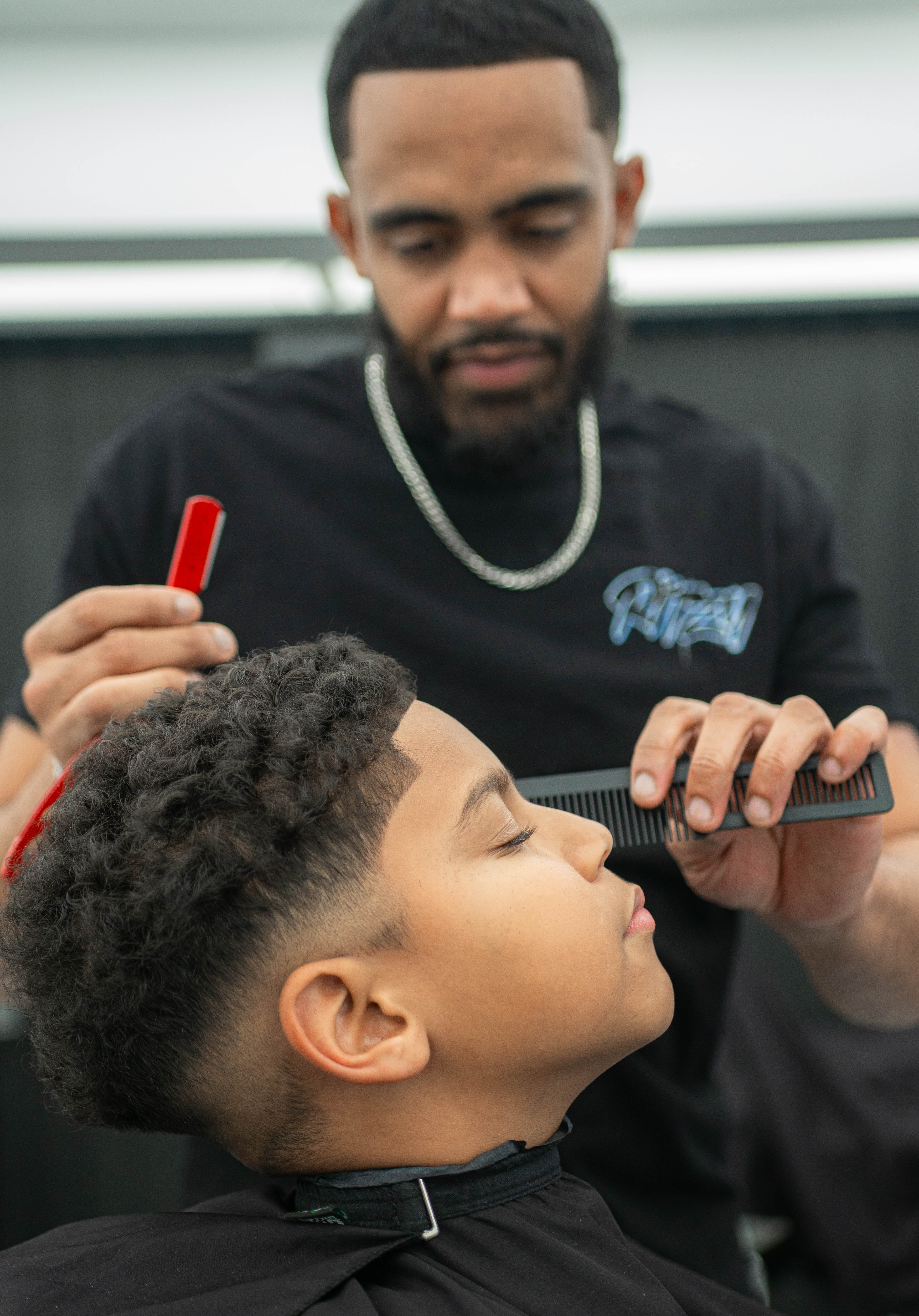 Barber carefully trimming a young boy's curly hair with a comb and clipper in a barbershop.