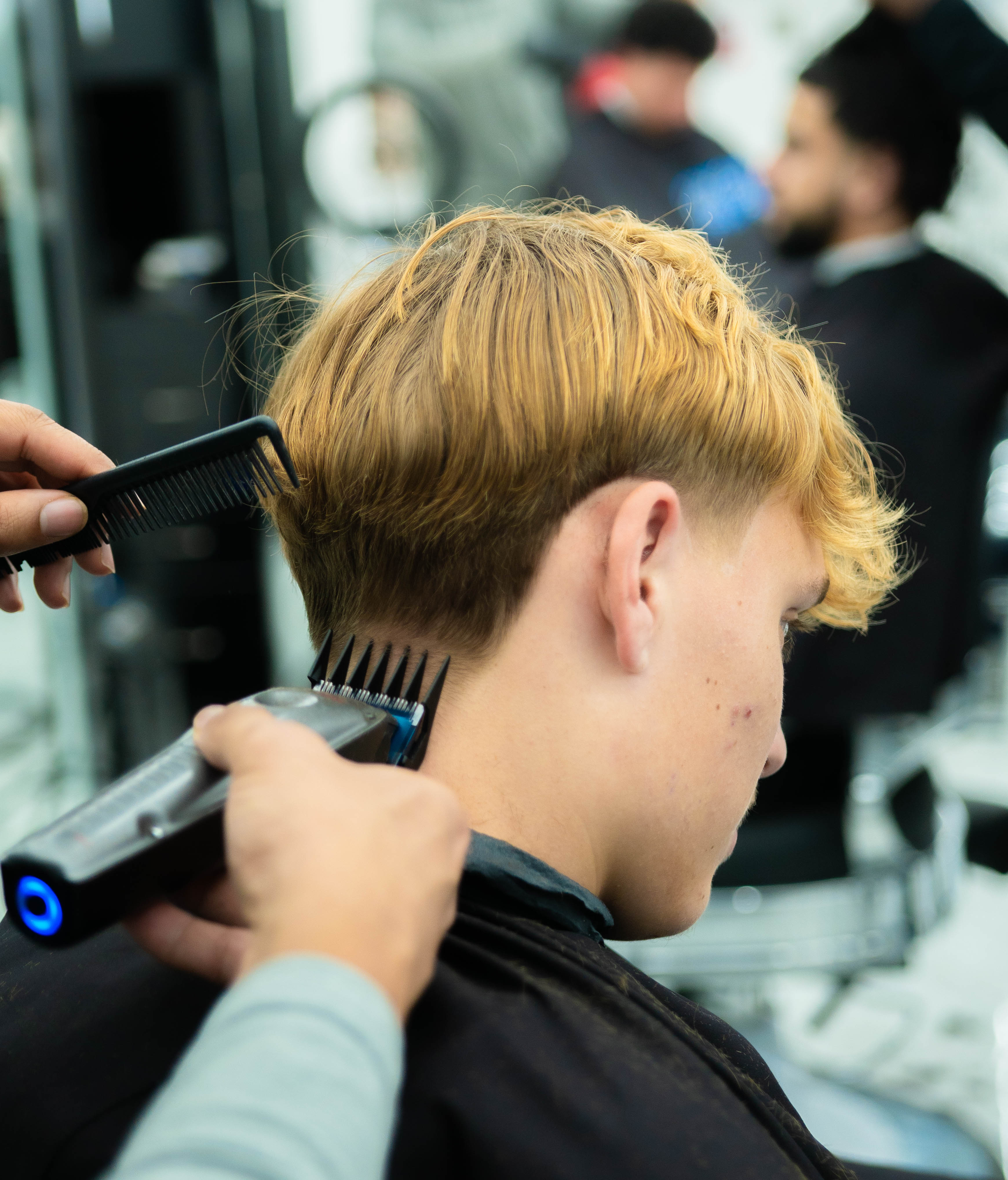 A barber using clippers and a comb to cut the back of a young man's blond hair in a salon.