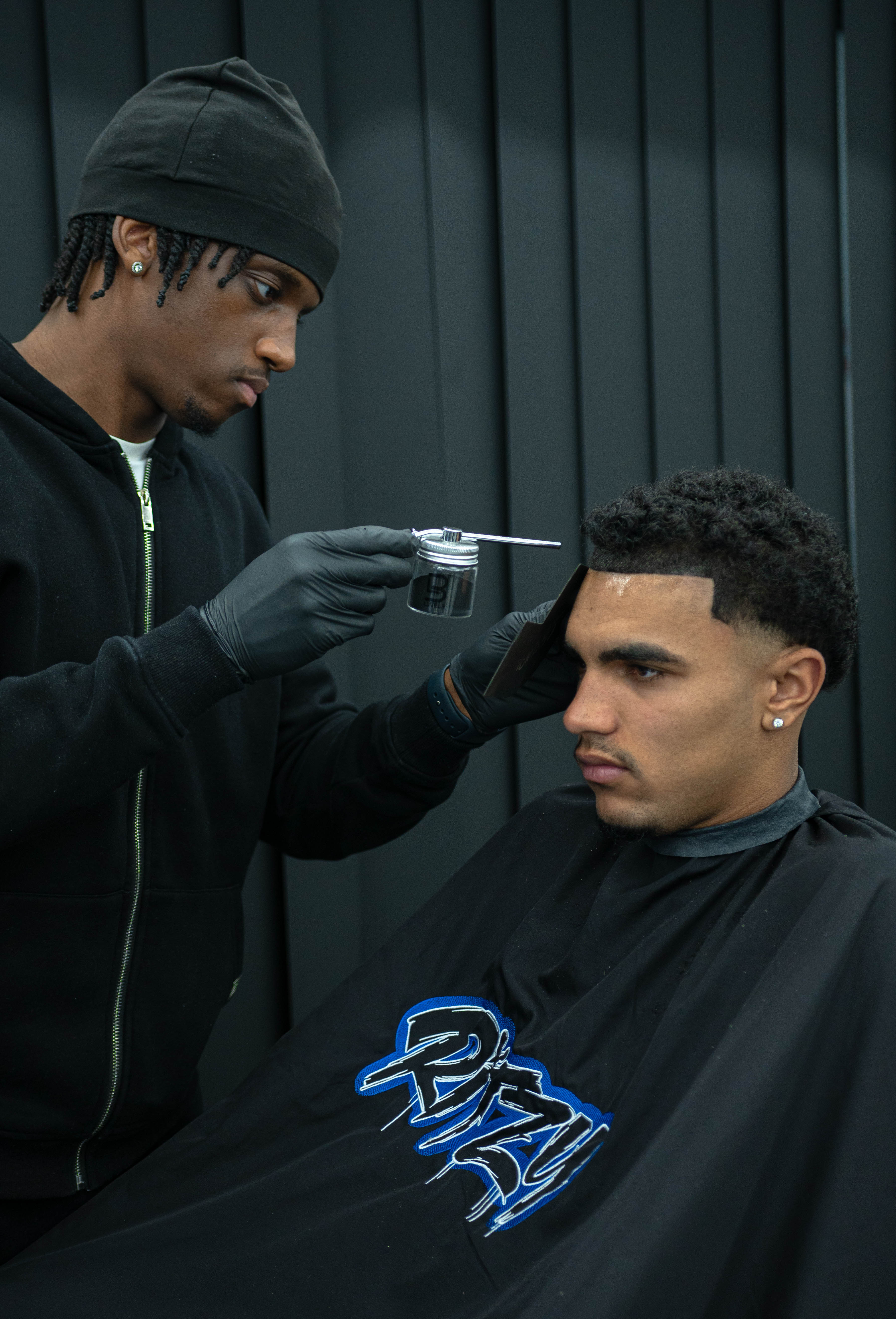 Barber wearing black gloves applies hair product to the front hairline of a young man seated under a black cape.
