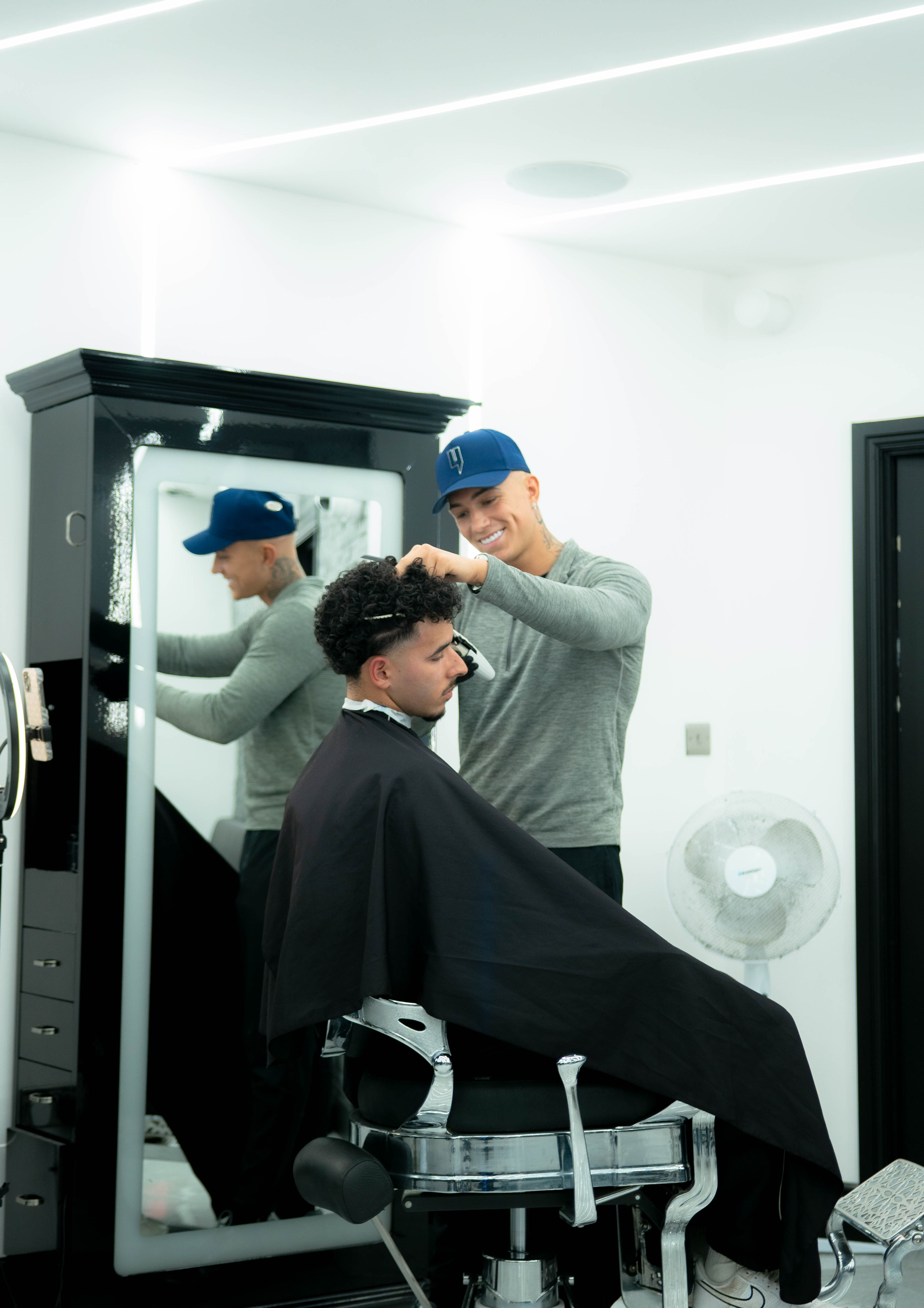 Barber wearing a blue cap cutting and styling a seated man's curly hair in a modern barbershop.
