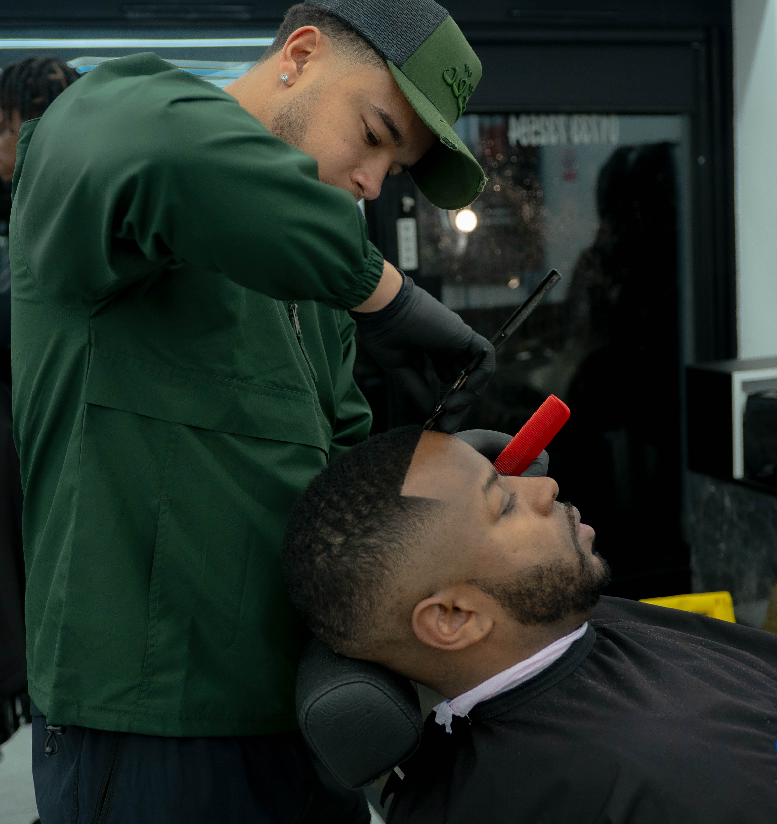 Barber in green jacket and cap carefully shaping a man's hairline with a razor in a barbershop  in Peterborough 