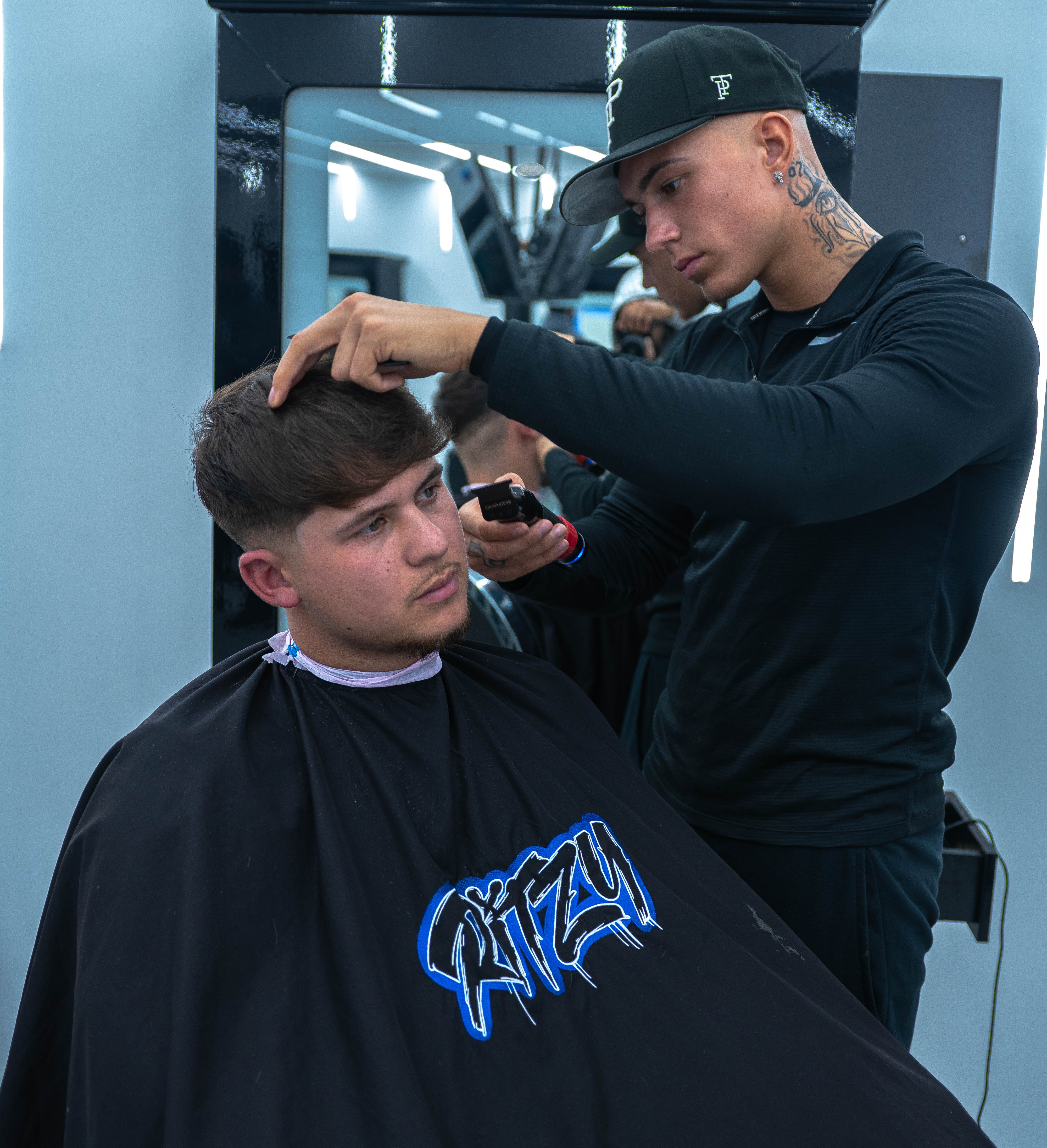 Barber wearing a black cap and shirt cutting a young man's hair in a modern barbershop.