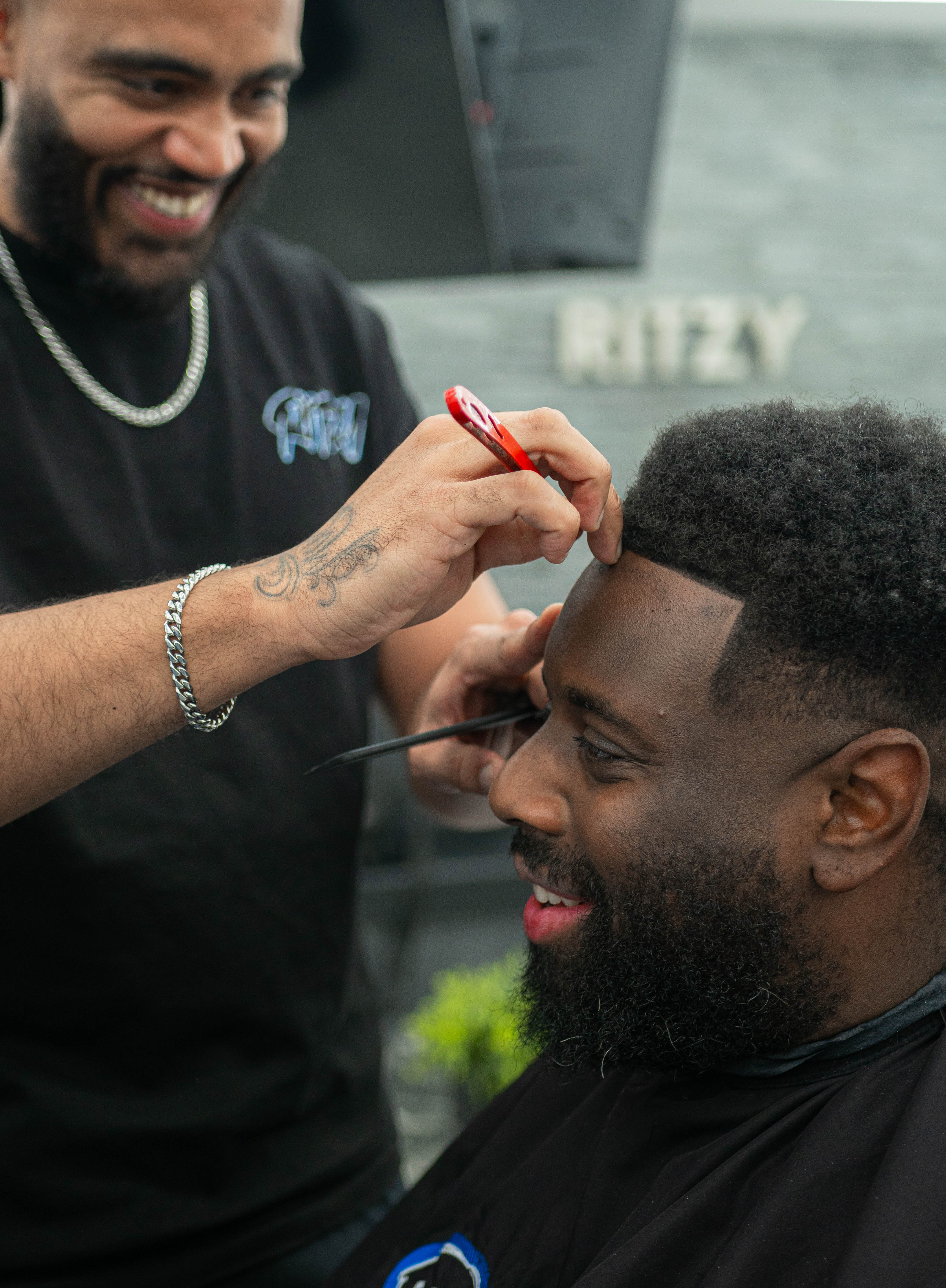 Barber smiling while trimming a man's hairline with a red razor in a barbershop.