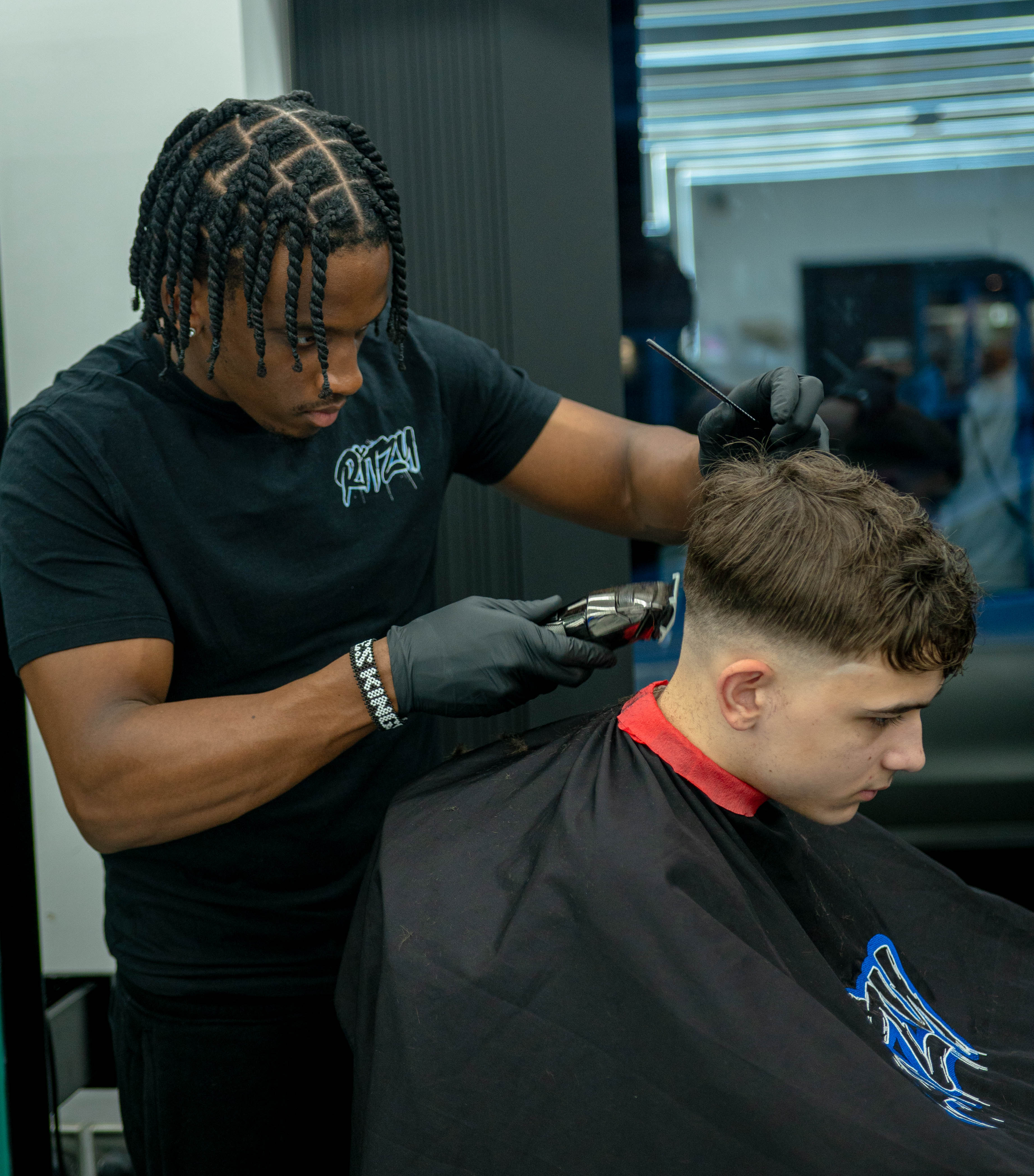 Barber with braided hair wearing black gloves trimming a young man's hair with clippers in a barbershop  in Peterborough 