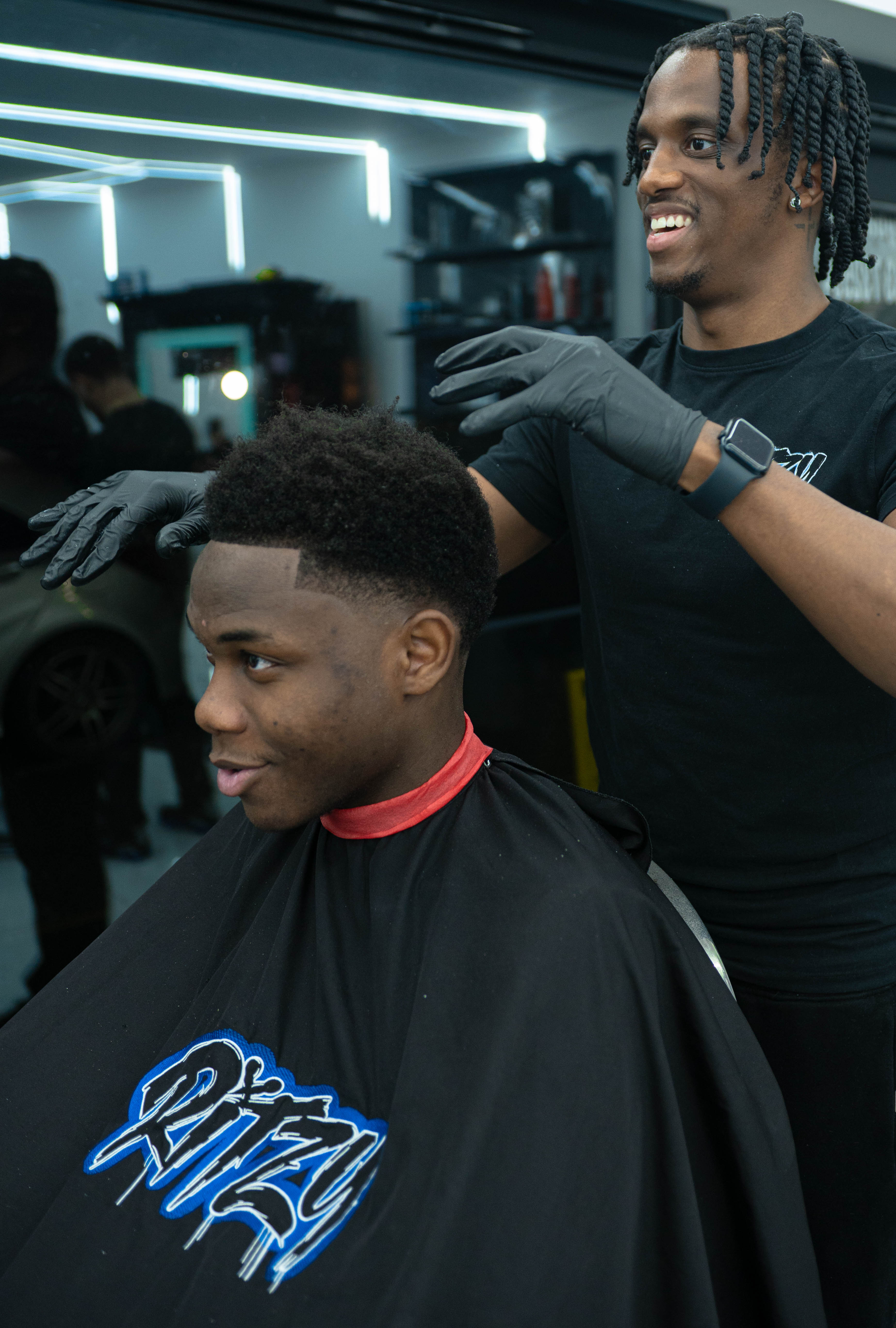 Barber with braided hair and black gloves styling a young man's short, faded haircut in a modern barbershop.
