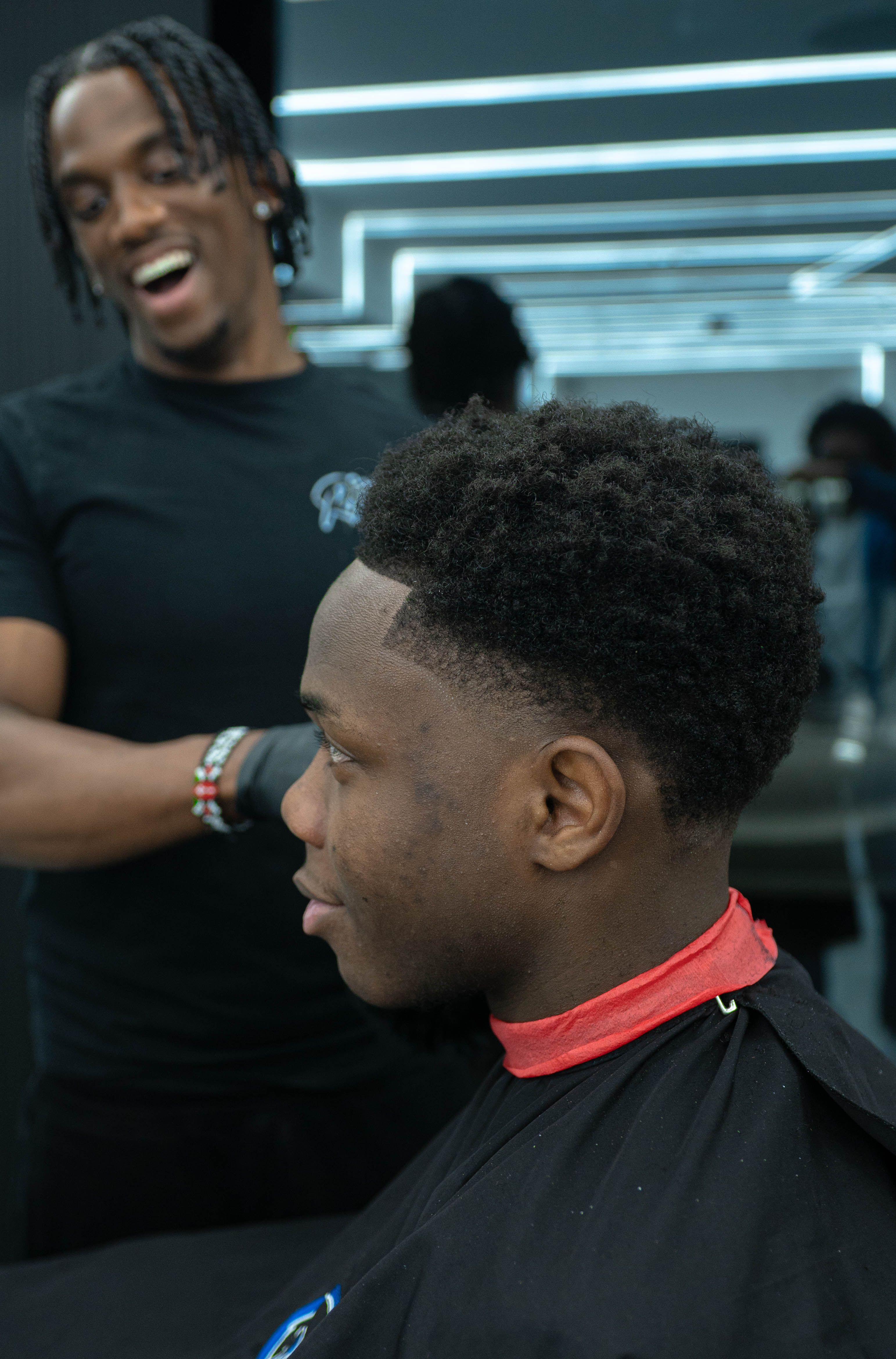 Young man with a fresh haircut sitting in a barbershop chair with a barber smiling behind him.