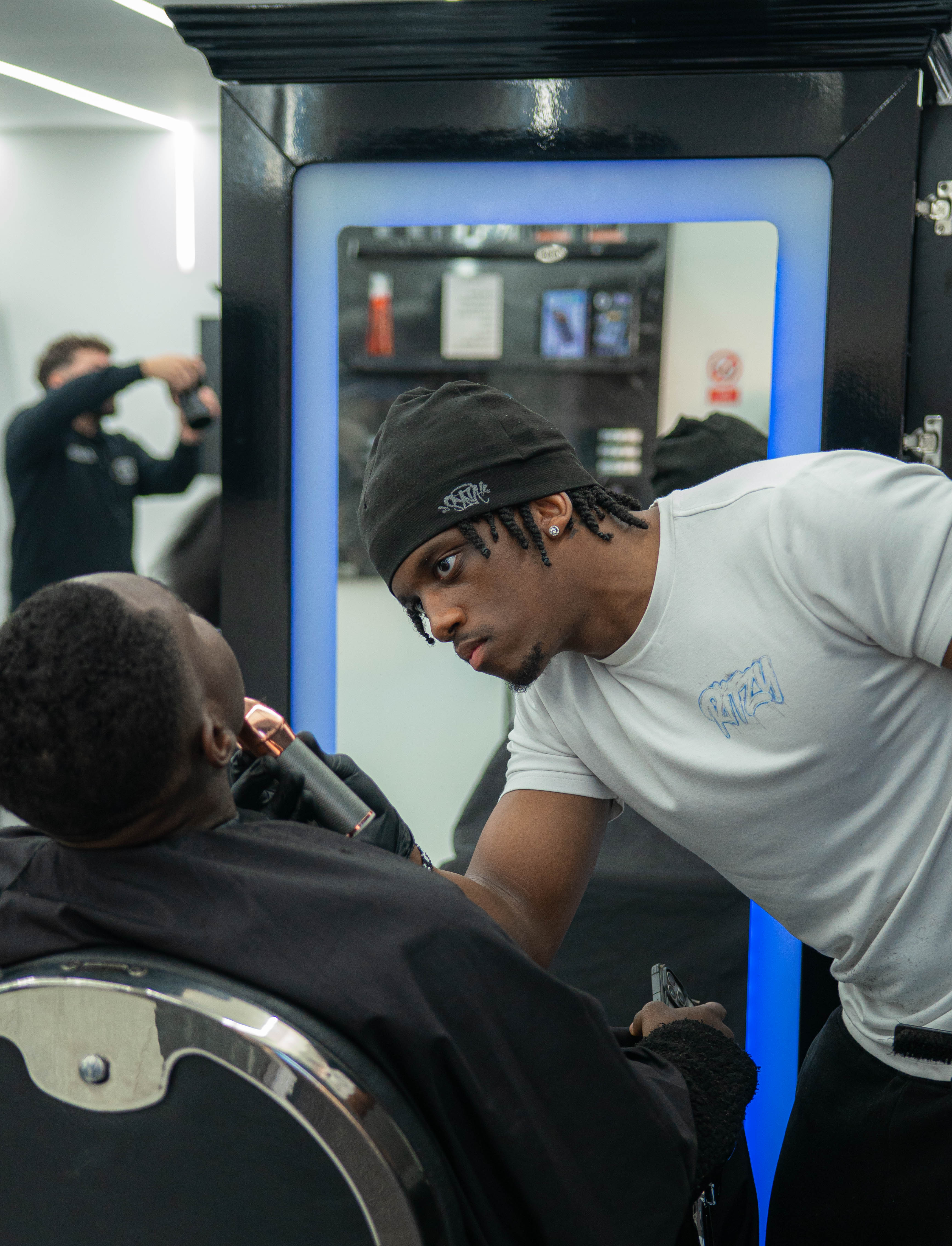 Barber wearing a black beanie and white shirt carefully trims a man's beard in a modern barbershop.