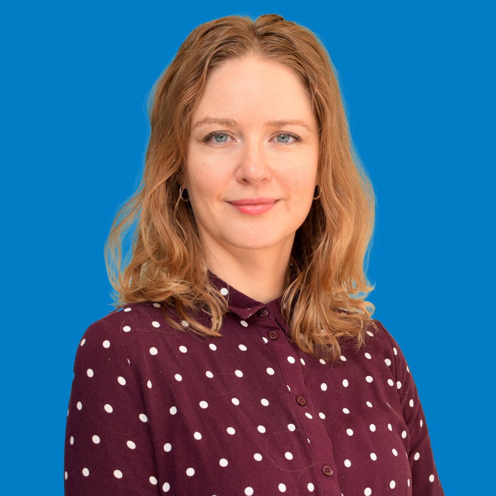 Portrait of a woman with shoulder-length wavy blonde hair wearing a maroon polka dot blouse against a blue background.