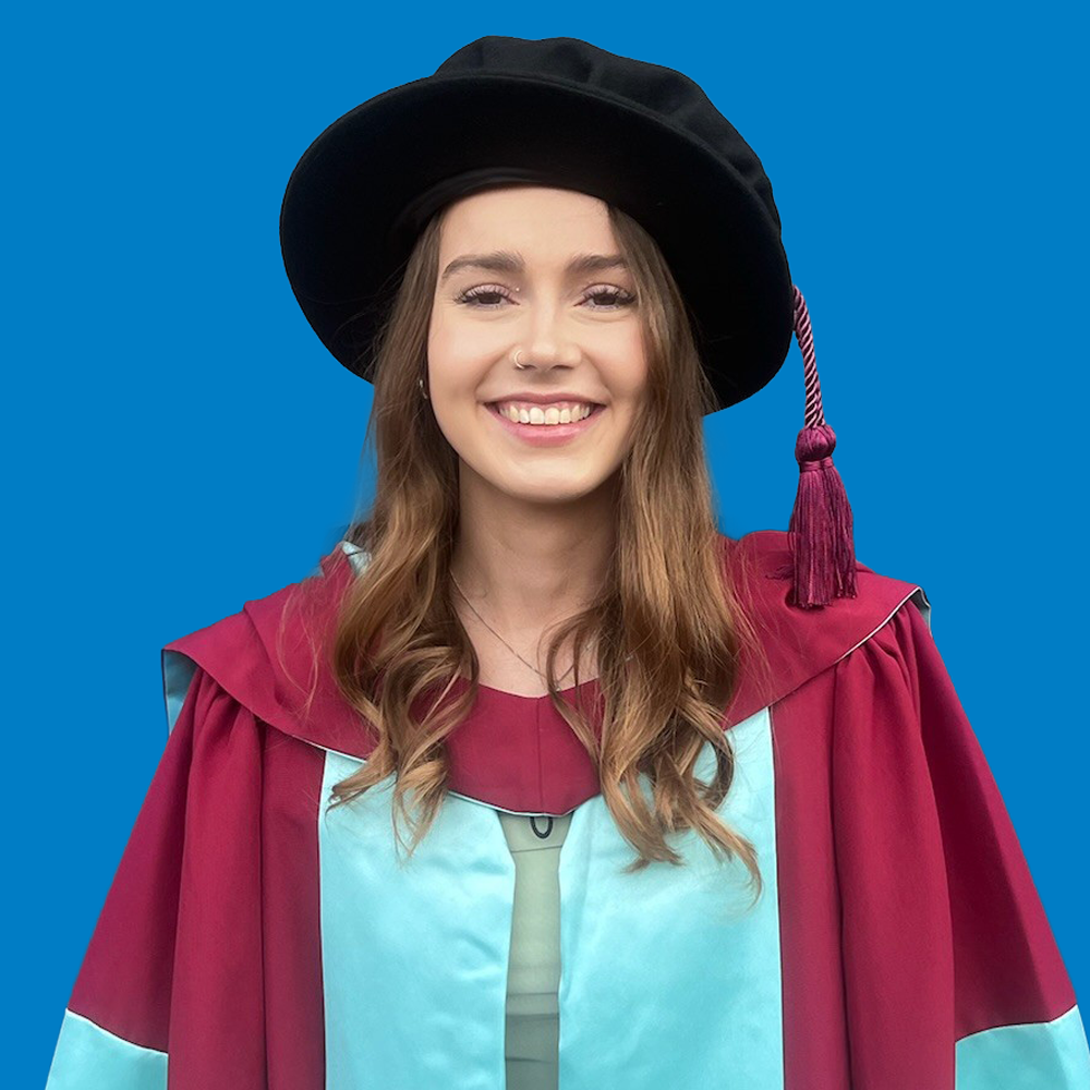 Smiling young woman wearing academic gown and black graduation cap with maroon tassel on blue background.