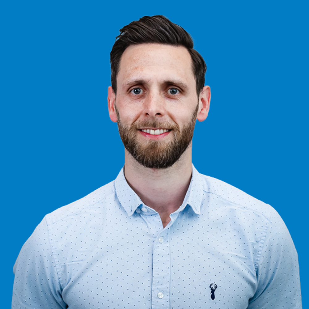 Smiling man with short dark hair and beard wearing a light blue shirt with small dots, against a blue background.