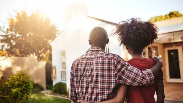 A view from behind as a man and woman stand side by side with their arms around one another in a yard while they appear to admire their home.