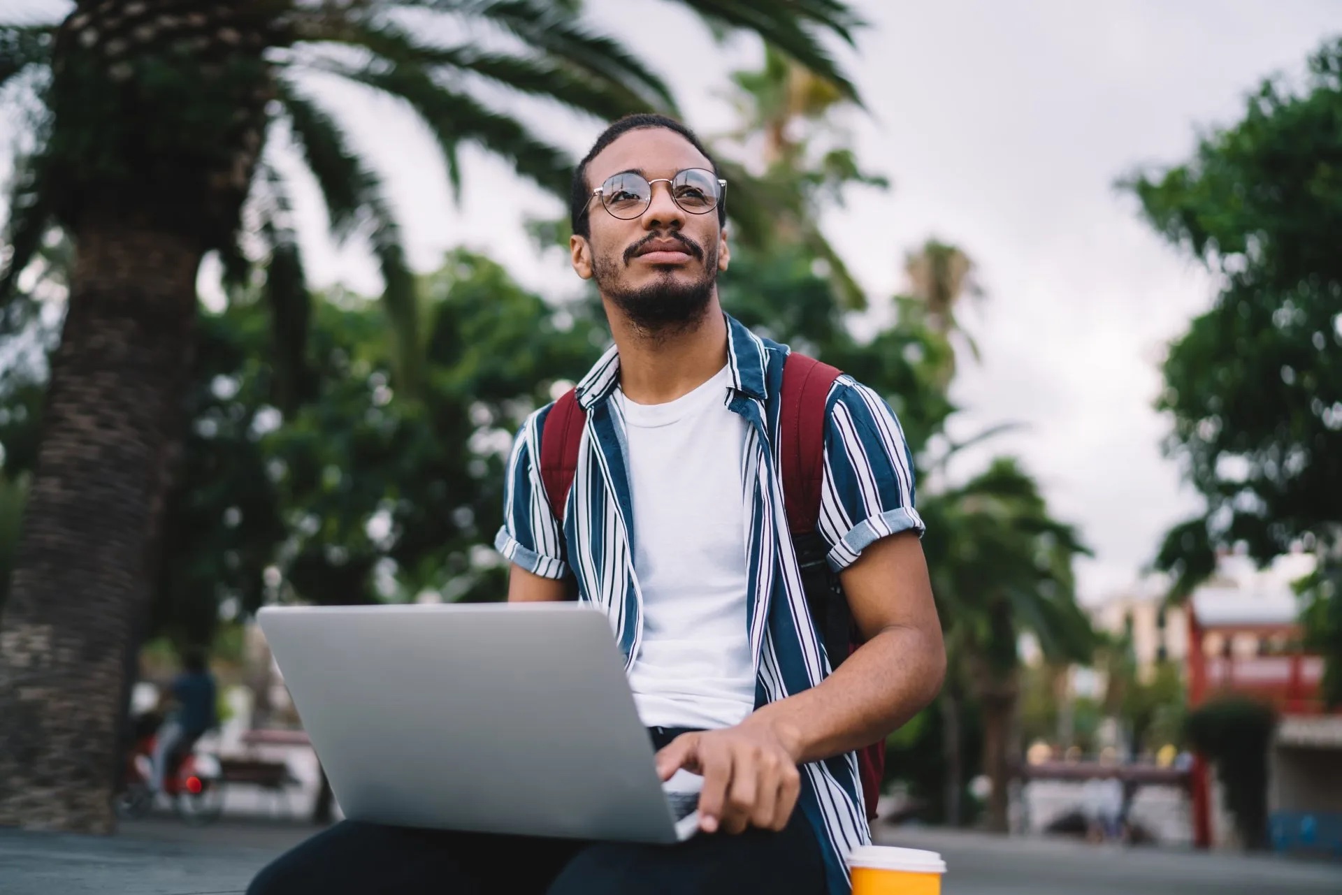 A man with glasses sporting a red backpack with a laptop in his lap looks on in the distance.