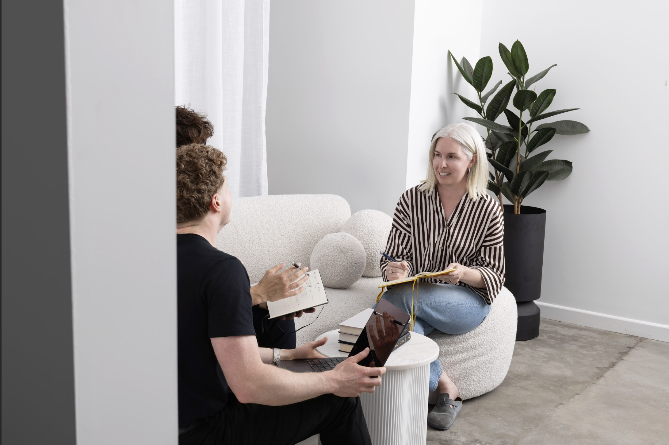 Three people engaged in a discussion in a modern, minimalist room with a potted plant, sitting on white curved furniture and holding notebooks and a laptop.