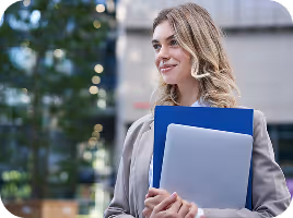 Smiling woman holding a blue folder and a laptop outdoors with trees and buildings in the background.