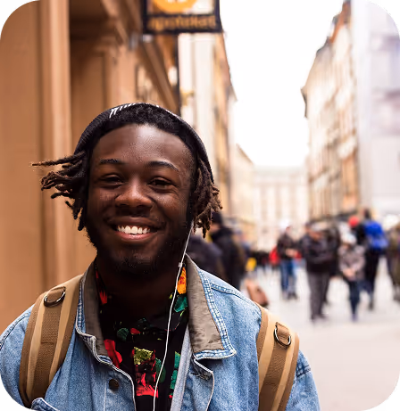 Smiling young man wearing a denim jacket, backpack, and earbuds stands in a busy urban street.