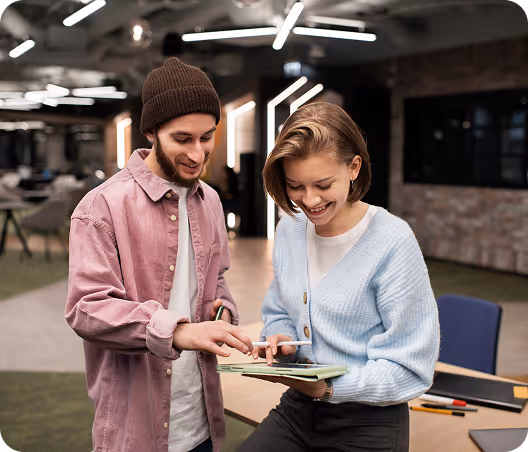 Two young adults smiling and discussing something on a tablet in a modern office setting.