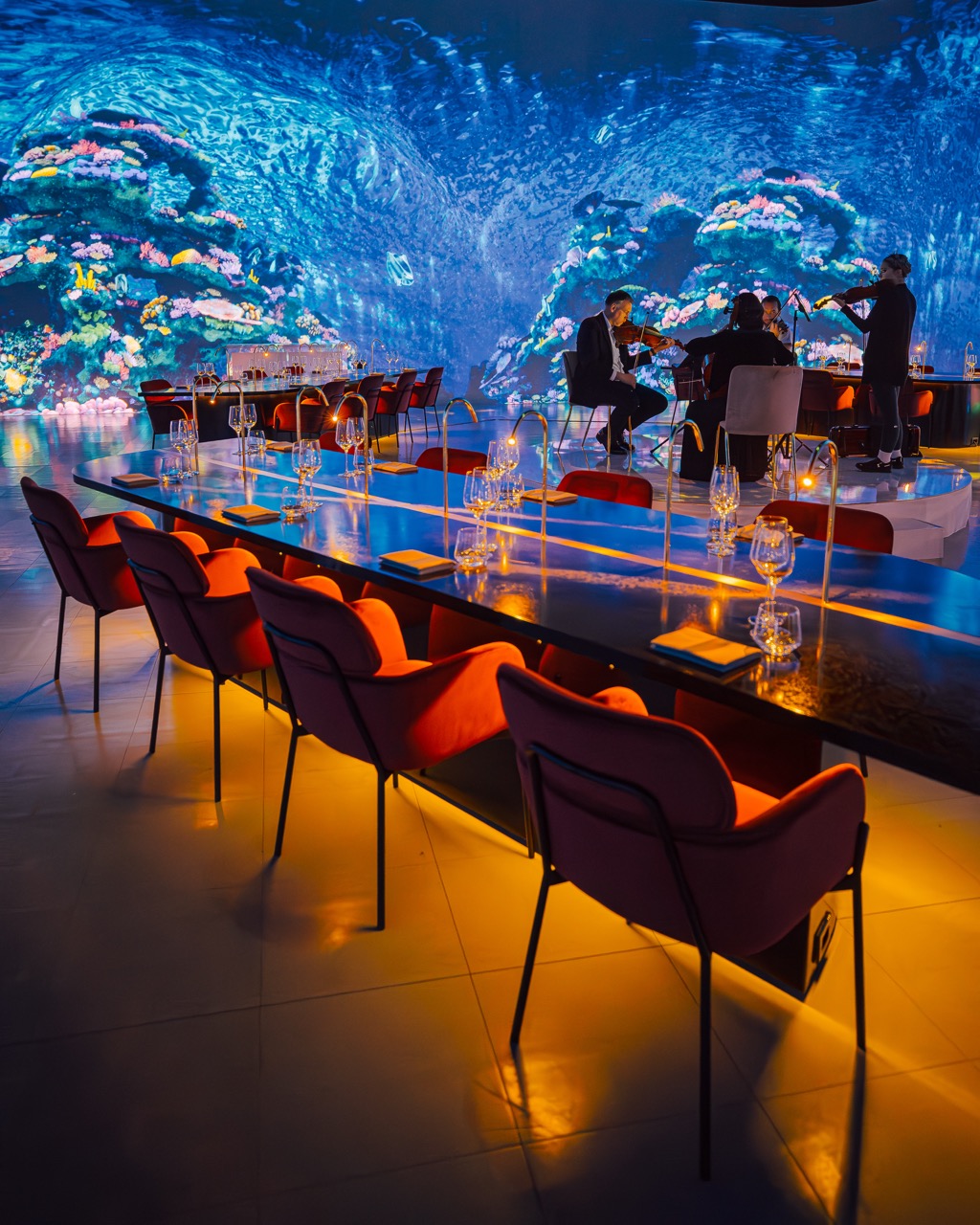 Dining setup with red chairs and a string quartet playing against an underwater coral backdrop projected onto curved walls for a luxury dinner event
