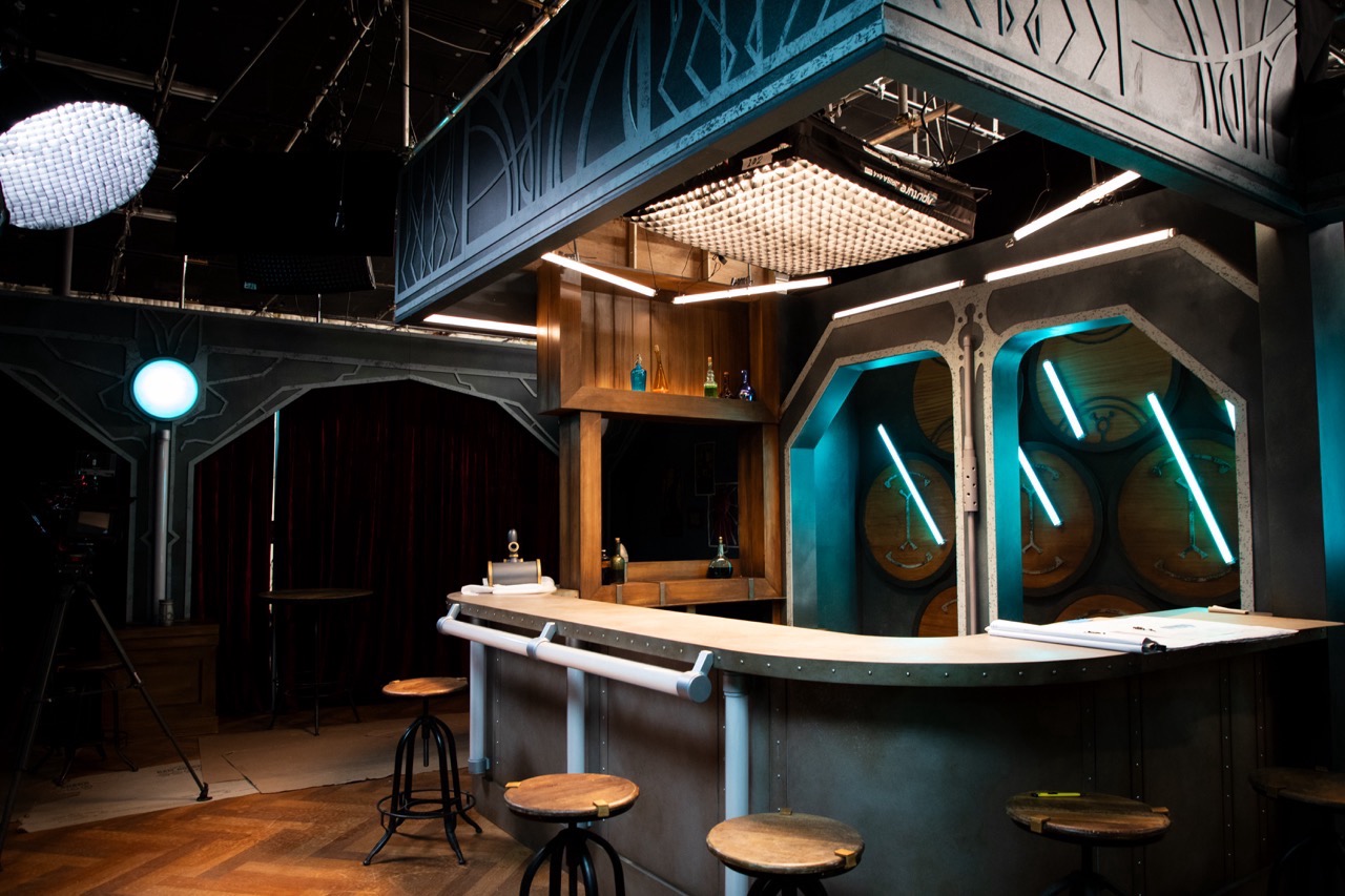Bar with wooden stools, neon lights, barrels, and cameras on a dimly lit set. It matches the bar from Arcane, and is used as a celebrity interview set.