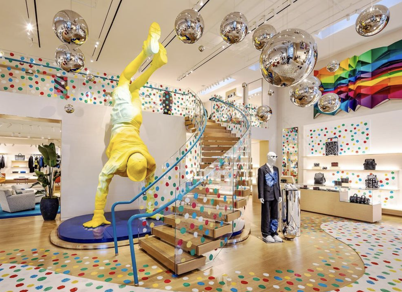 Retail space with colorful polka dot decor, staircase with glass railing, silver hanging spheres, and mannequins.