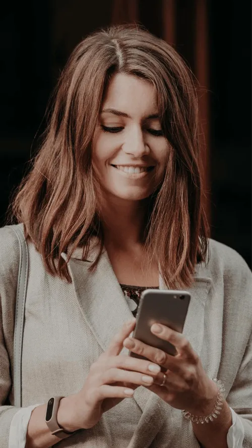 Smiling woman with shoulder-length brown hair wearing a beige coat, looking at her smartphone.