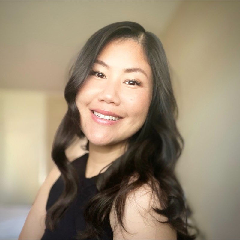 Smiling woman with long wavy black hair wearing a sleeveless black top against a neutral background.