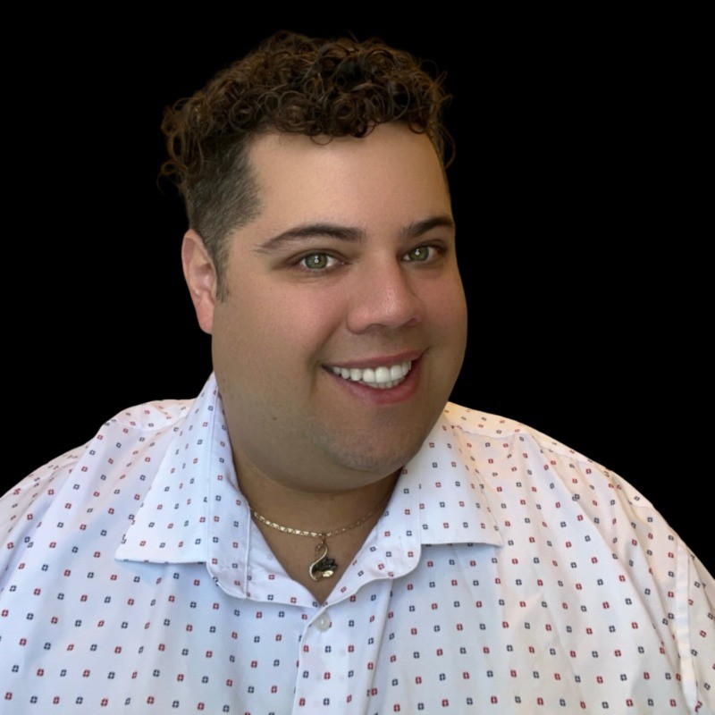 Smiling man with curly dark hair wearing a white patterned shirt and a necklace with a pendant against a black background.