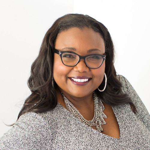 Smiling woman with dark hair, wearing glasses, hoop earrings, a silver chain necklace, and a textured gray top against a white background.