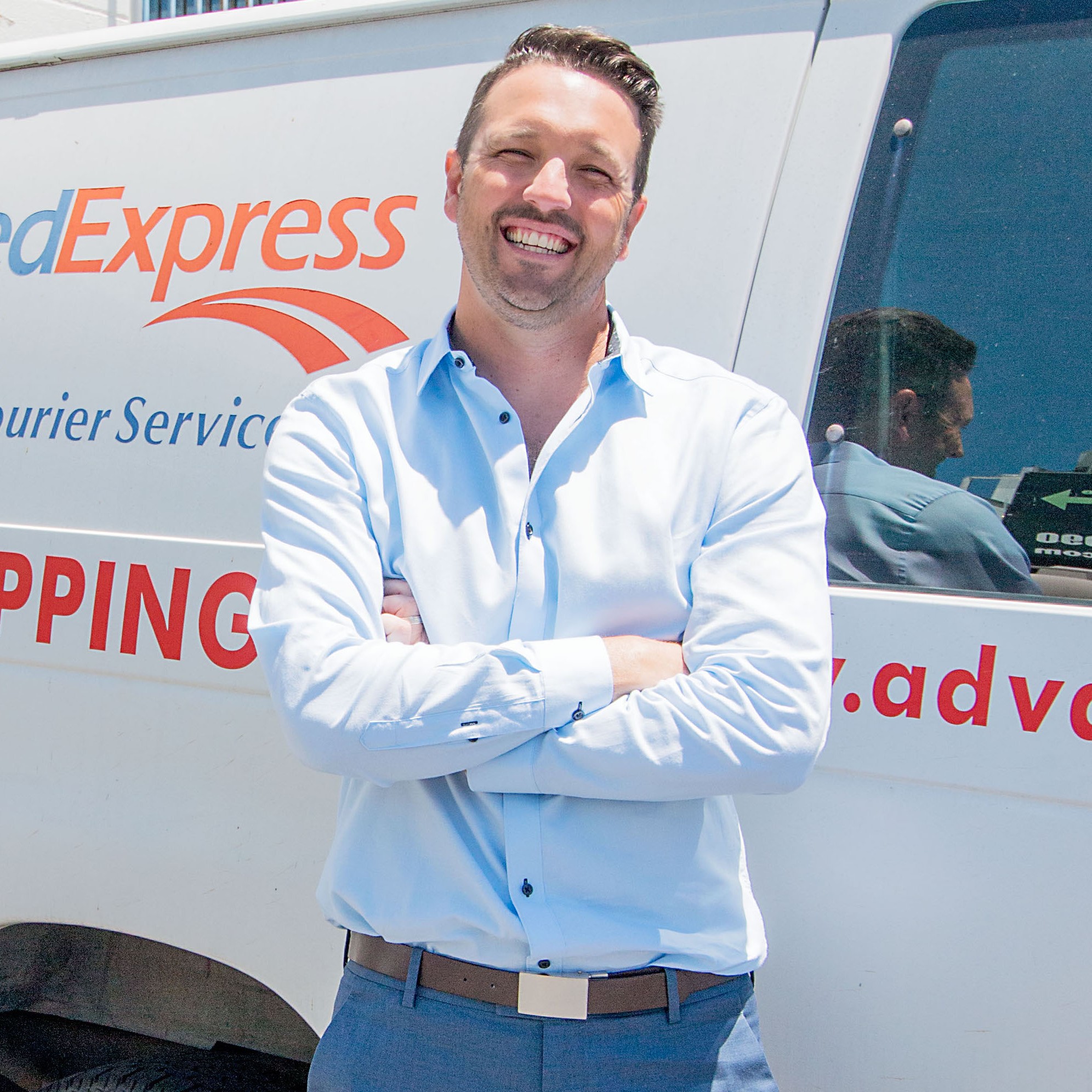 Smiling man in light blue shirt with arms crossed standing in front of a FedExpress delivery van.
