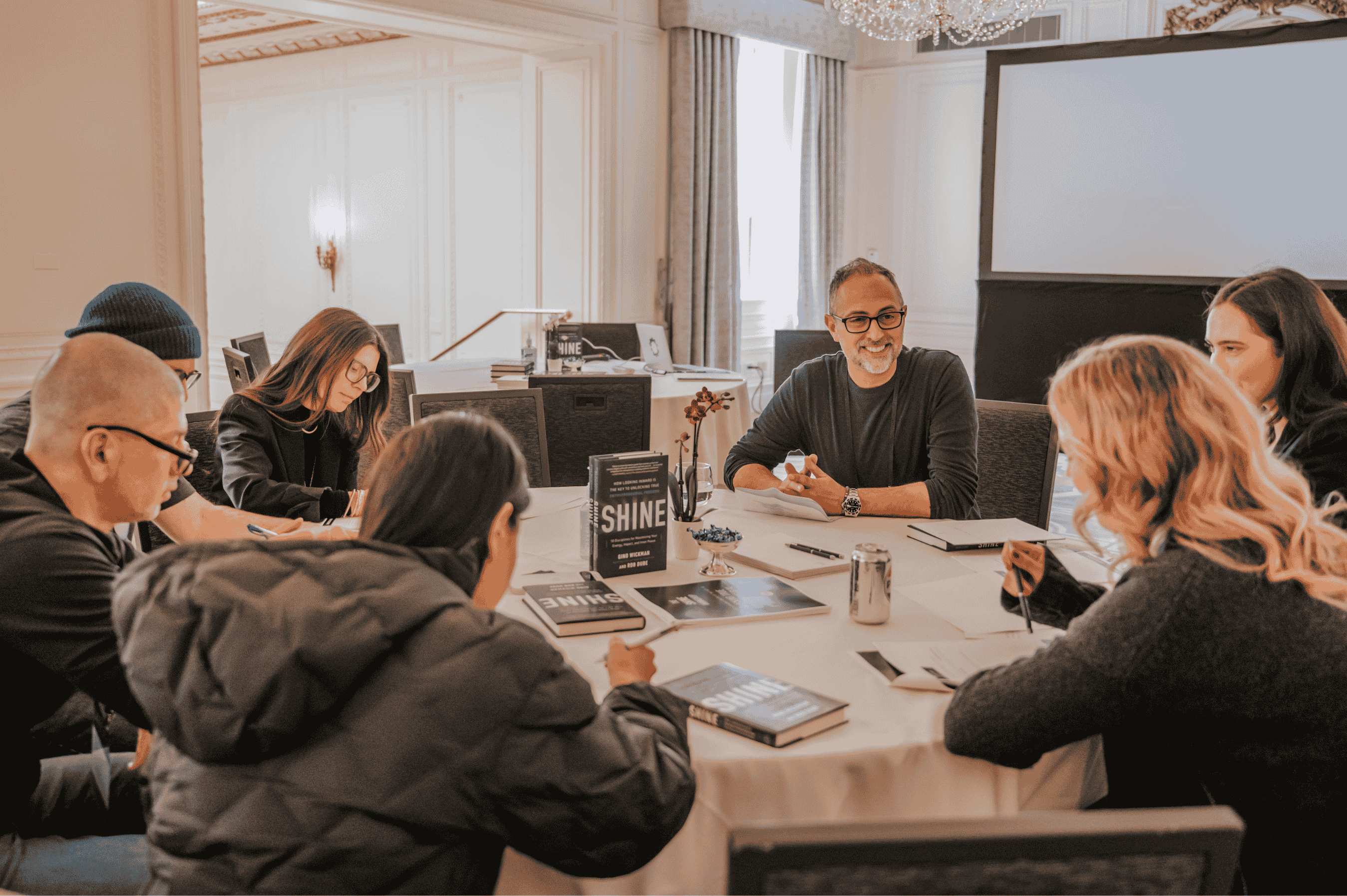 Group of six people sitting around a table in a meeting room, discussing with books titled 'SHINE' on the table, lead by Philip Pfeifer.