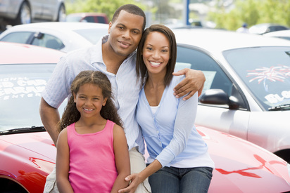 Smiling African American family of three sitting on the hood of a red car in a parking lot.