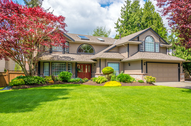 Two-story suburban house with beige siding, solar panels on the roof, large green lawn, and trees with red and green leaves.