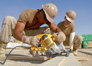 Two construction workers wearing hard hats and gloves operate a circular saw cutting wood outdoors under a clear blue sky.