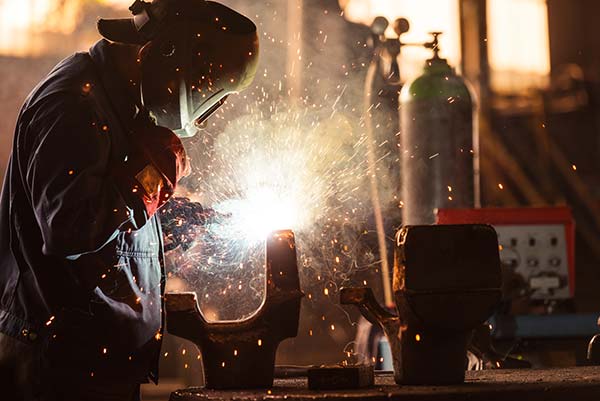 Person wearing welding gear working with sparks and bright light from welding in an industrial workshop.