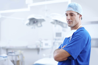 Male healthcare professional in blue scrubs and hairnet standing with arms crossed in a bright medical facility.