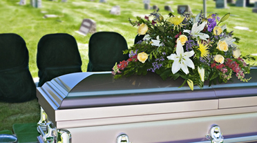 Casket with a floral arrangement on top at a graveside service with empty black-covered chairs in the background.
