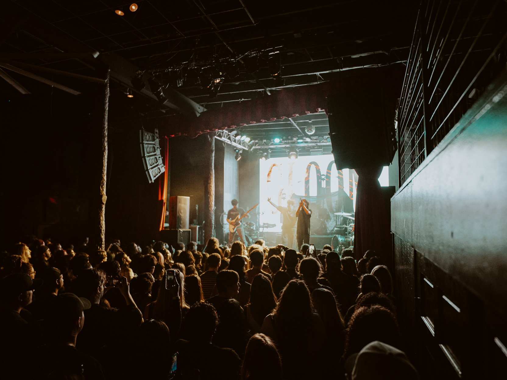 Crowd watching a live band performing on a dimly lit stage at an indoor concert venue.