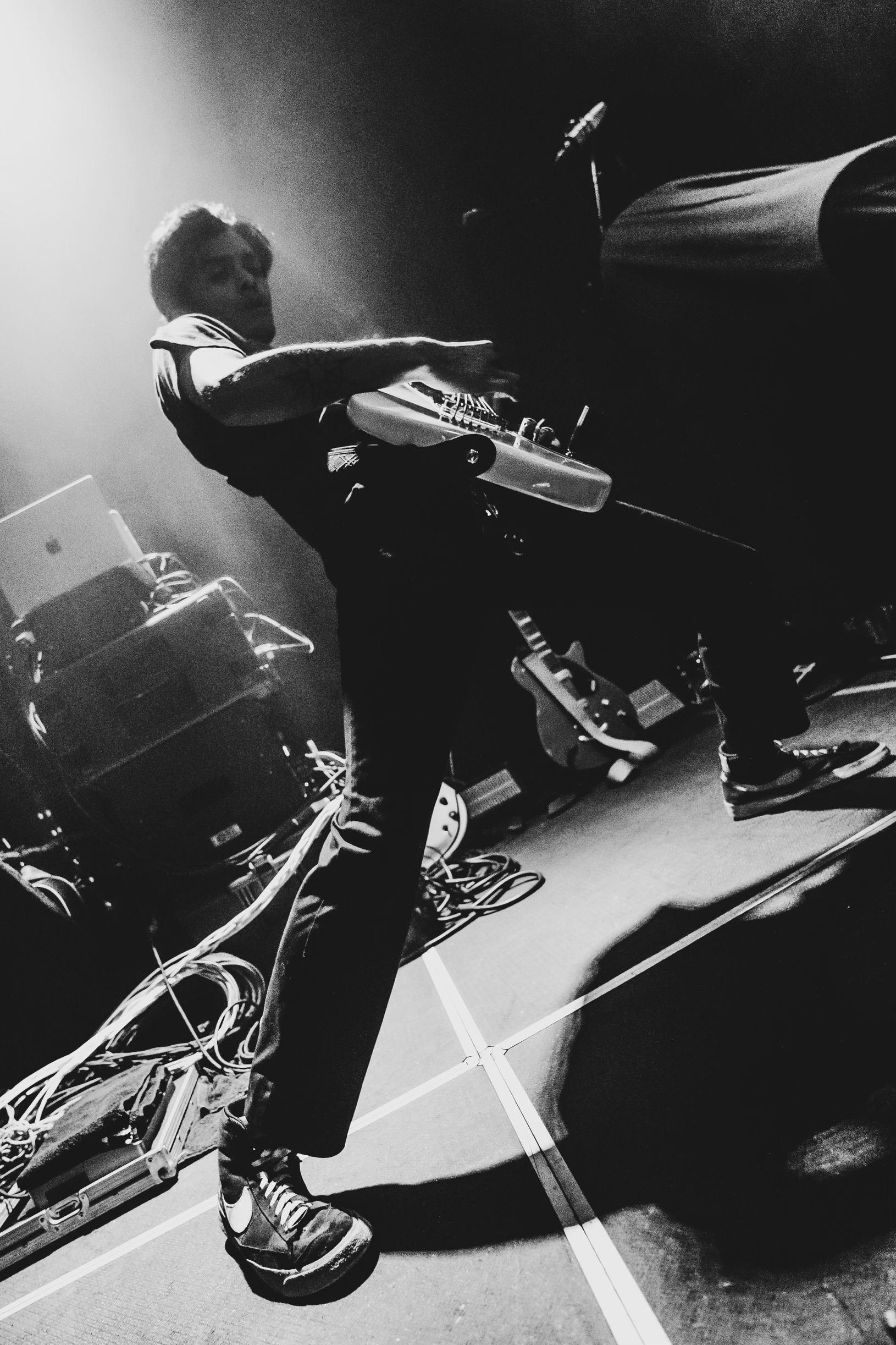 Black and white photo of guitarist Aaron Medina from gloom leaning back while playing an electric guitar on stage with cables and another guitar in the background.