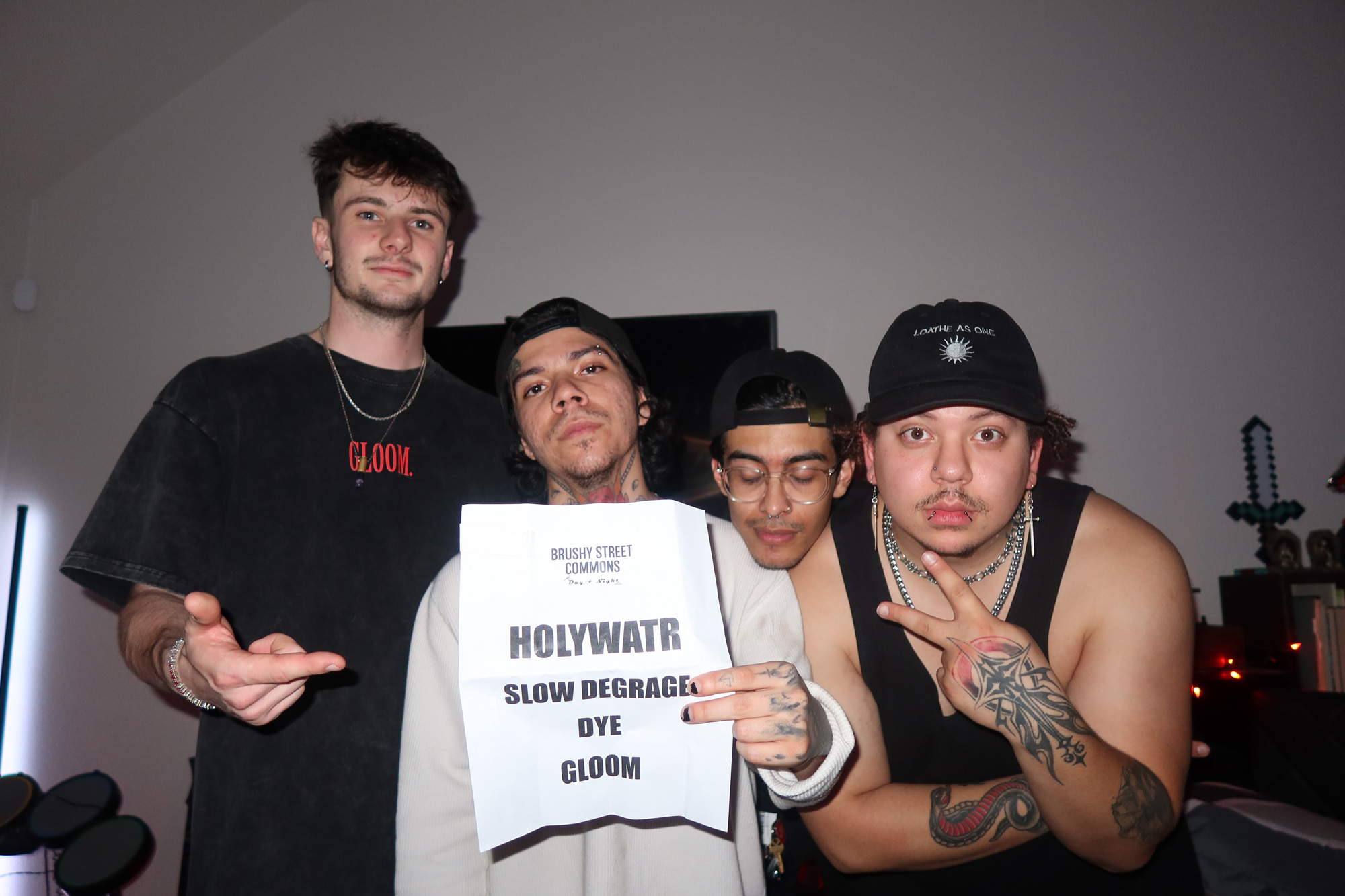 Gloom posing indoors, one holding the lineup of their event including HOLYWATR, SLOW DEGRADE, DYE, and GLOOM.