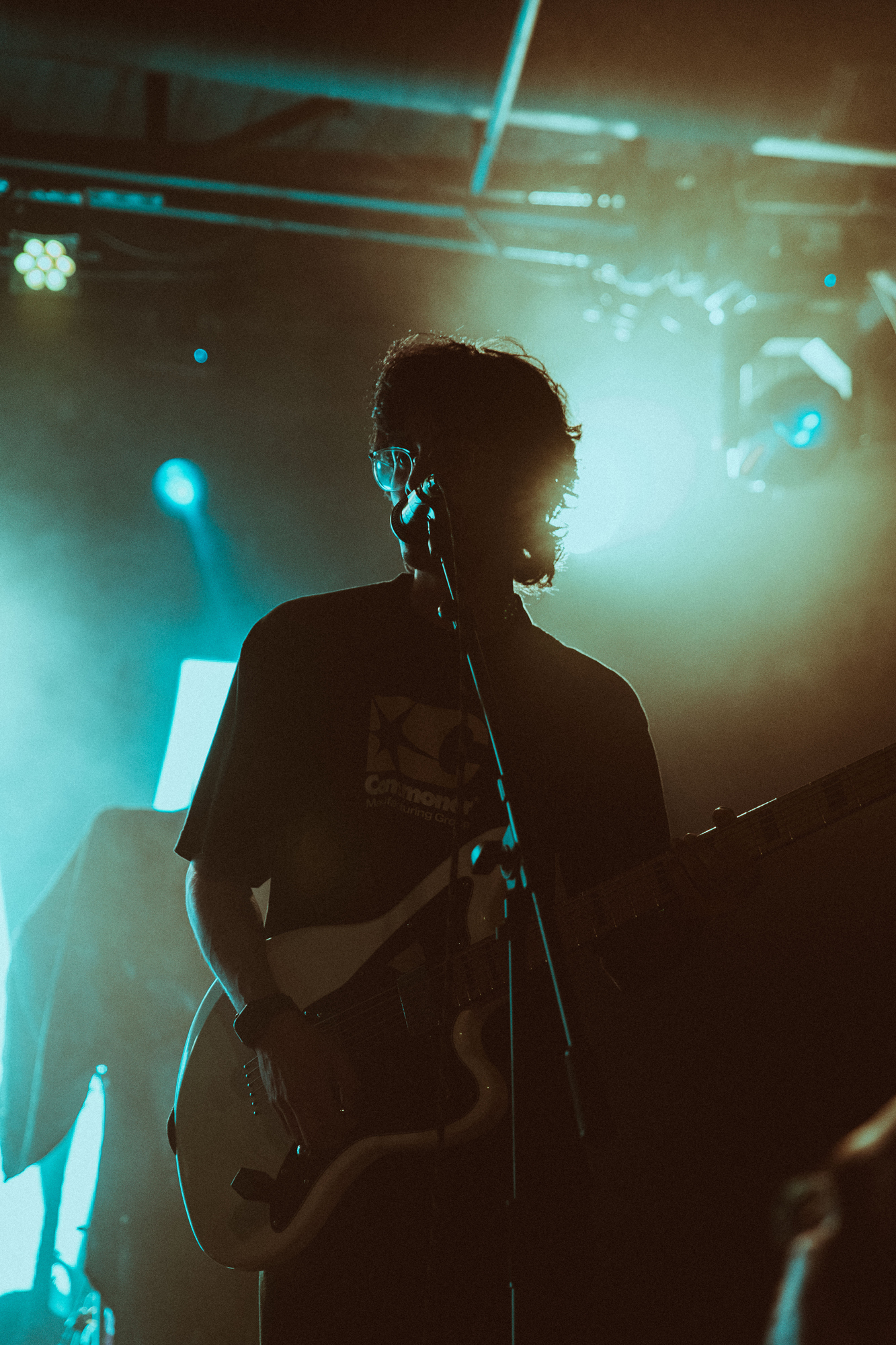 Silhouetted musician wearing glasses playing electric guitar with microphone in front under stage lights.