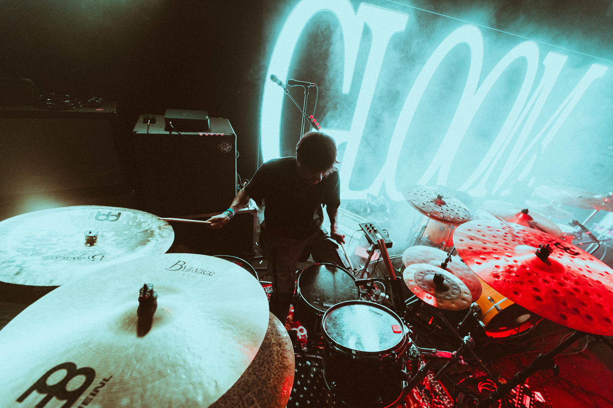 Drummer playing a drum set on stage with bright neon lights and fog in the background.