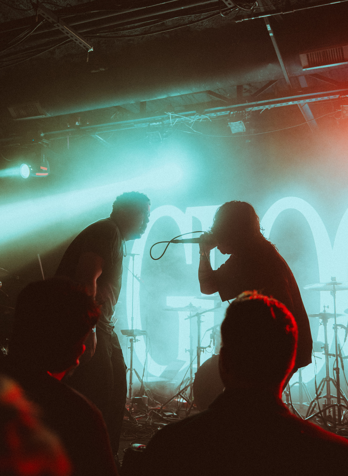 Silhouettes of two performers on stage with microphone and drum set under blue and red lighting.