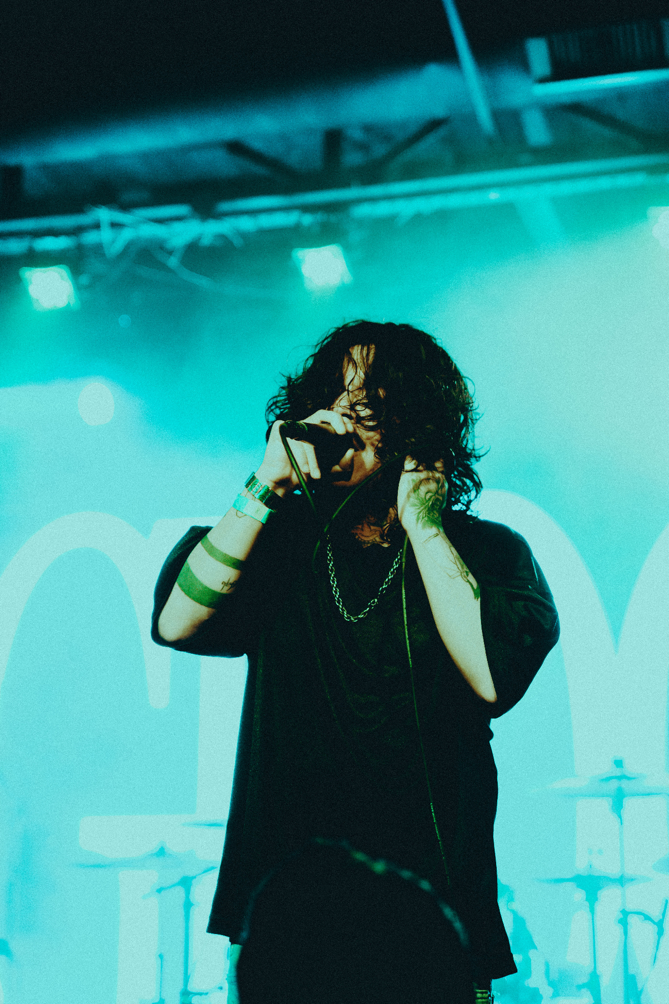 Male singer with curly hair holding a microphone and performing on stage under teal lighting.