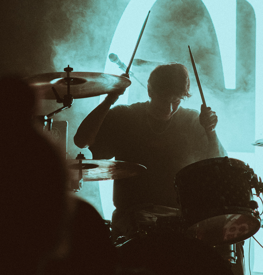 Silhouette of a drummer playing drums with sticks raised in a smoky, blue-lit setting.