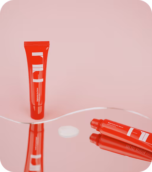 Two red cosmetic tubes on a reflective pink surface, one standing upright and the other lying down with the cap removed.