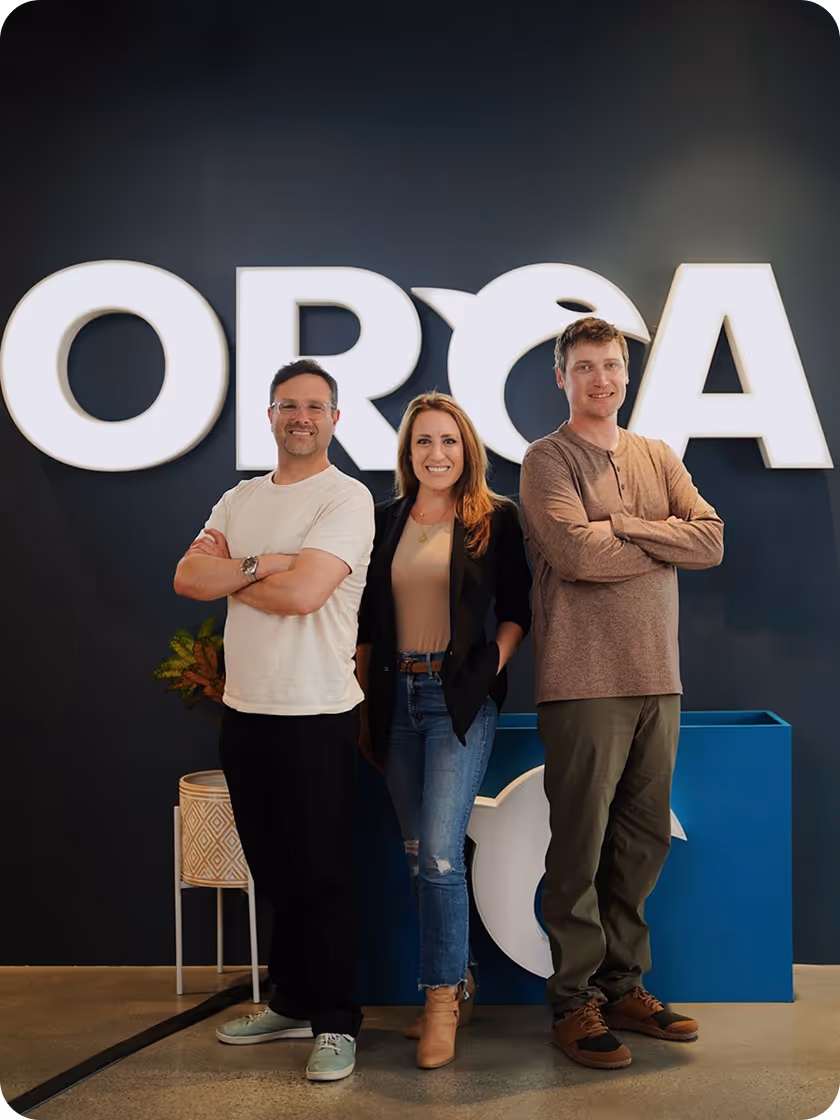 Three smiling adults standing in front of a wall with a large illuminated ORCA sign, two men with arms crossed on either side of a woman with hands in pockets.