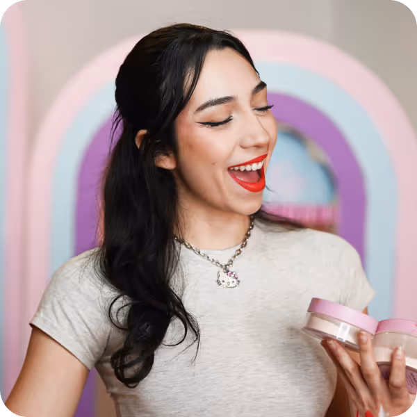 Smiling woman with long dark hair wearing red lipstick and a Hello Kitty necklace, holding two cosmetic containers.