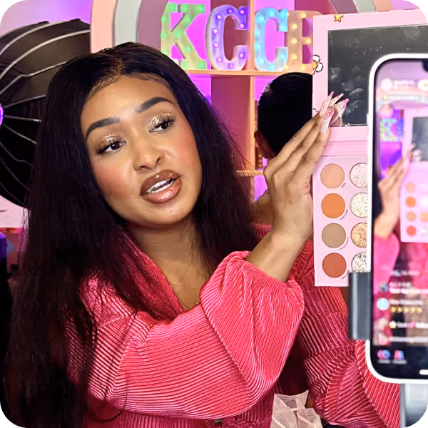 Woman in a pink top demonstrating a makeup palette in front of a smartphone camera during a live video.