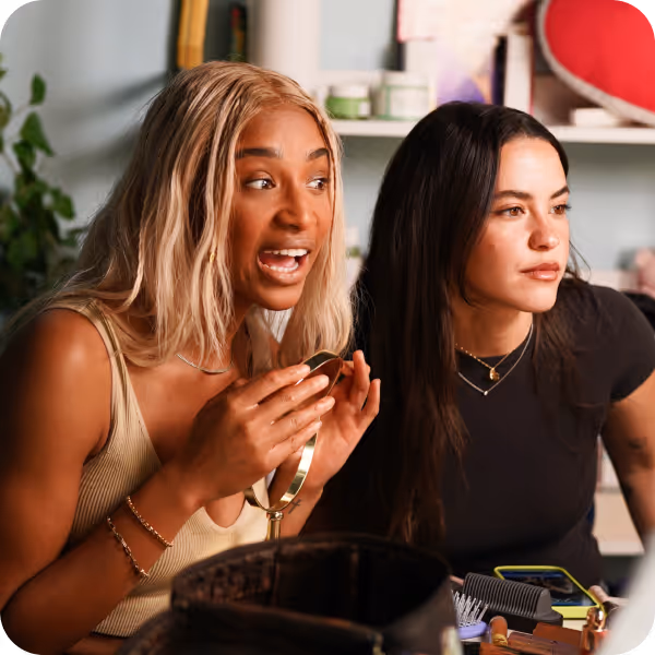 Two women sitting at a table with makeup tools, one holding a small mirror while talking, the other looking ahead.